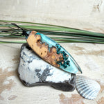 Handgefertigter, länglicher Surfbrett-Anhänger aus naturbelassenem Maserholz und transparentem Harz in Lagunen-Türkis, an schwarzem Lederband mit Knoten – maritimer Unikatschmuck für Damen und Herren, dekorativ mit Stein und Muschel inszeniert.