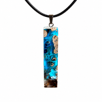 Rechteckiger Stabanhänger aus Maserholz und glasklarem Epoxidharz in tiefen Blau- und Türkistönen mit organischer Lagunen/Riff-Optik. Hochglanzpoliert, ca. 5,3 × 1,2 cm. Der Anhänger hängt an einer schwarzen Lederkette mit Edelstahl-Karabiner und 5-cm-Verlängerung (45 + 5 cm); die Anhängerschlaufe ist ebenfalls aus Edelstahl. Handgefertigtes Unikat, Lieferung in edler Schmuckbox – maritimer Look.