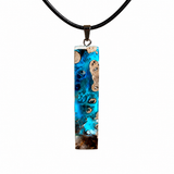 Rechteckiger Stabanhänger aus Maserholz und glasklarem Epoxidharz in tiefen Blau- und Türkistönen mit organischer Lagunen/Riff-Optik. Hochglanzpoliert, ca. 5,3 × 1,2 cm. Der Anhänger hängt an einer schwarzen Lederkette mit Edelstahl-Karabiner und 5-cm-Verlängerung (45 + 5 cm); die Anhängerschlaufe ist ebenfalls aus Edelstahl. Handgefertigtes Unikat, Lieferung in edler Schmuckbox – maritimer Look.