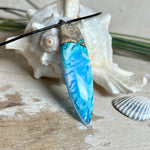 Surfbrett-Anhänger aus Maserholz und blau-weißem Epoxidharz, hochglanzpoliert, ca. 6,5 × 2 cm. An schwarzer Lederkette mit Edelstahl-Karabiner und 5-cm-Verlängerung; Edelstahl-Öse. Im Tageslicht lagunenblau, unter Schwarzlicht fluoreszierend – ideal als Party-/Festival-Schmuck. Handgefertigtes Unikat, Lieferung in edler Schmuckbox.