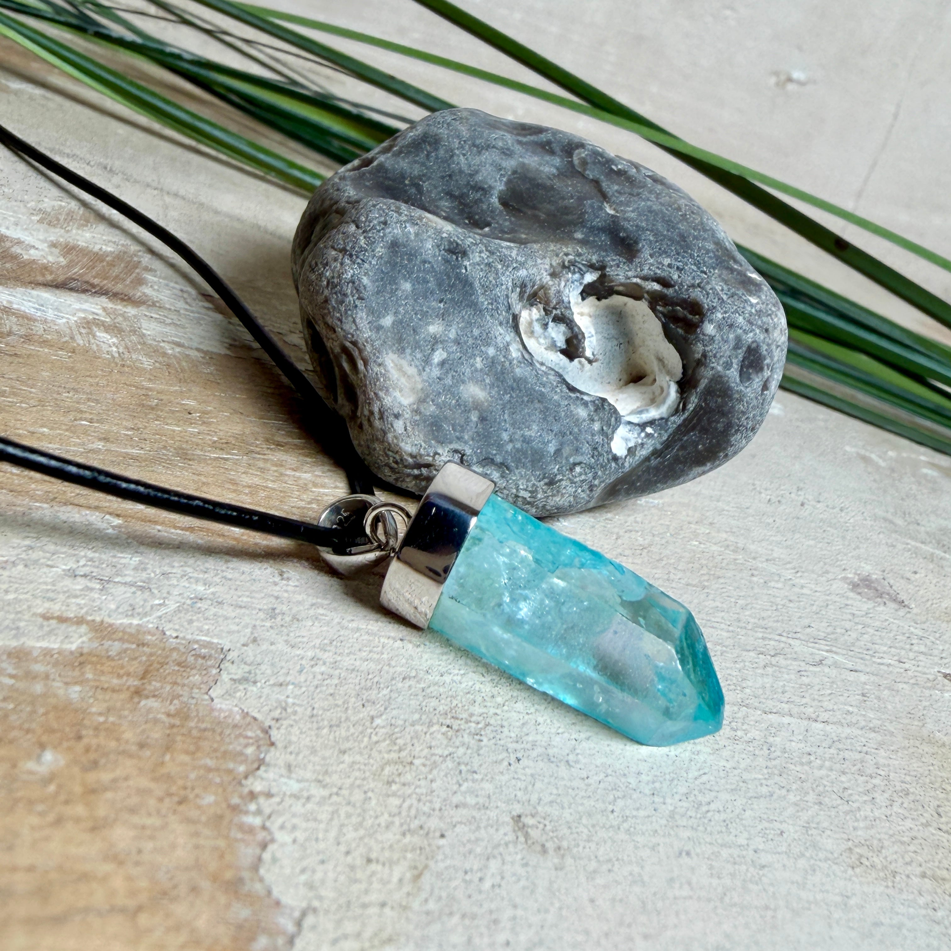 Nahaufnahme einer handgefertigten Halskette mit Aqua Aura Rohkristall: naturbelassener, sechskantiger Bergkristall mit goldbedampfter Aqua-Beschichtung in Türkisblau, gefasst in 925er Silber und getragen an einer schwarzen Lederkette mit Edelstahl-Karabiner und Verlängerungskettchen.