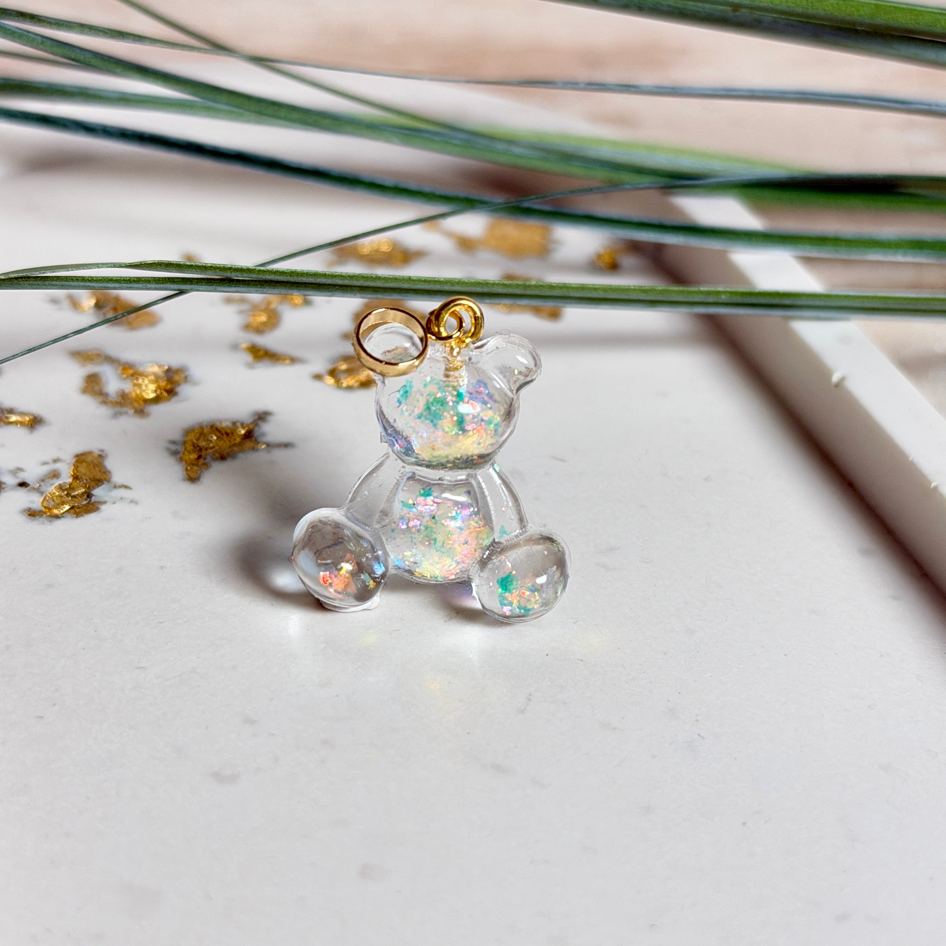 Kleiner Bärchen-Anhänger aus transparentem Harz mit irisierenden Flakes in Regenbogen-Schimmer, goldfarbene Anhängerschlaufe aus Edelstahl. Größe ca. 2,2 × 2,1 cm. Handgefertigtes Unikat; Flakes-Anordnung variiert, vereinzelt kleine Luftbläschen möglich.