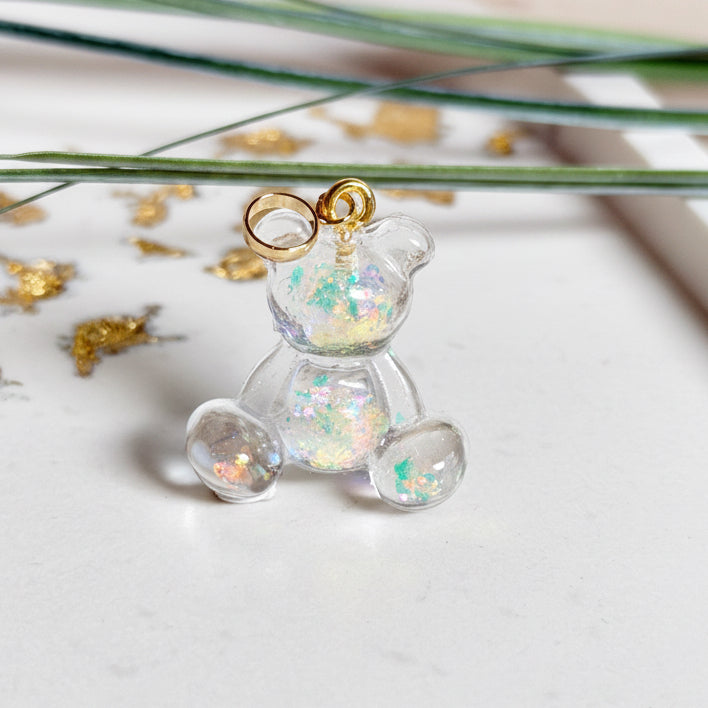 Kleiner Bärchen-Anhänger aus transparentem Harz mit irisierenden Flakes in Regenbogen-Schimmer, goldfarbene Anhängerschlaufe aus Edelstahl. Größe ca. 2,2 × 2,1 cm. Handgefertigtes Unikat; Flakes-Anordnung variiert, vereinzelt kleine Luftbläschen möglich.