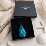 Tropfenanhänger aus Maserholz und türkis-blauem Epoxidharz mit weißer Gischtoptik am schwarzen Lederband, präsentiert in schwarzer Schmuckbox von ZeitlosSchmuckDesign – handgefertigter maritimer Naturschmuck.