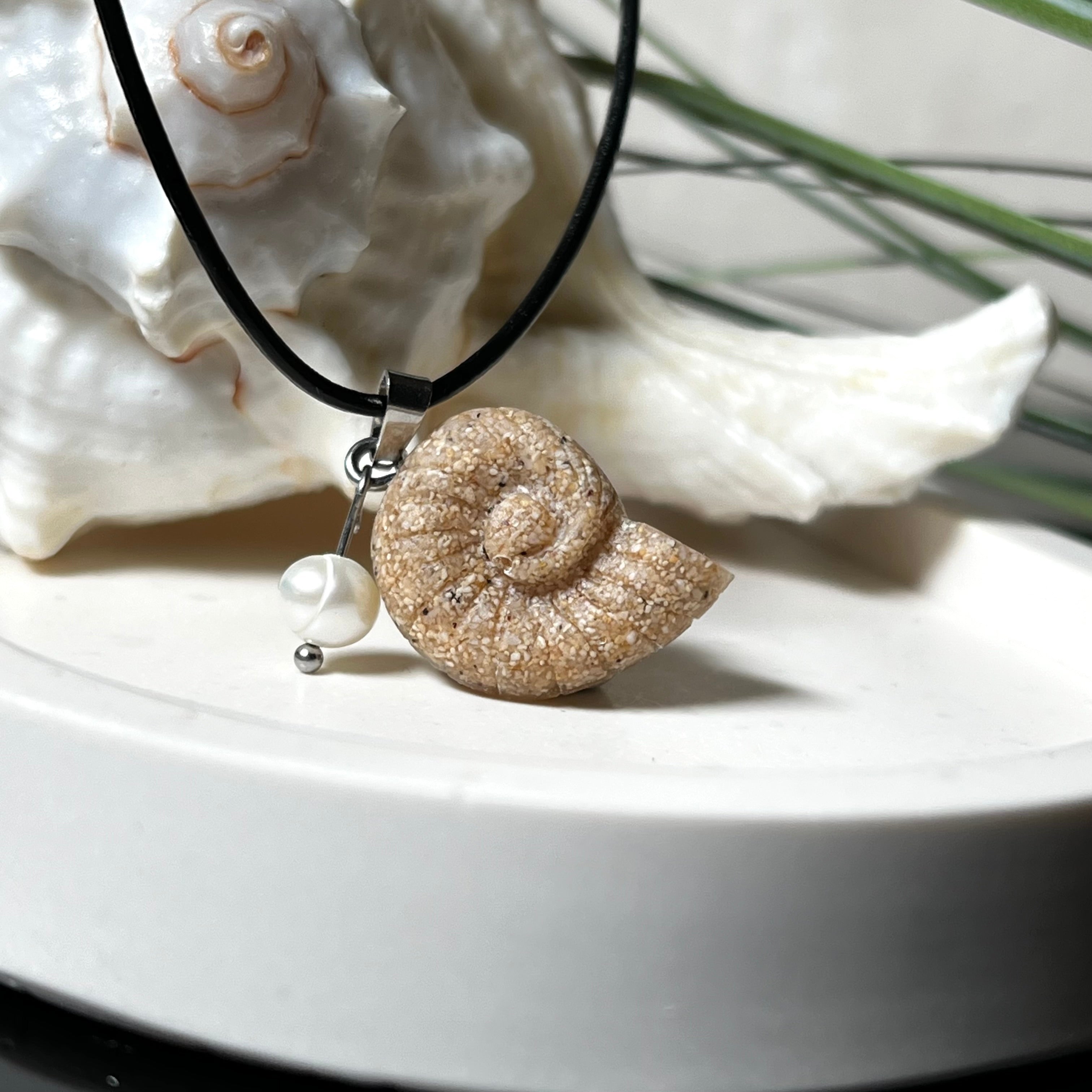 Handgefertigter Ammonit-Anhänger aus echtem Meersand & Harz mit filigraner Edelstahlöse und einer edlen Süßwasserperle. Maritimer Schmuck, inspiriert von der Kraft und Schönheit des Ozeans. Jedes Stück ein Unikat. Präsentiert auf einer Muschel, mit Lederkette.