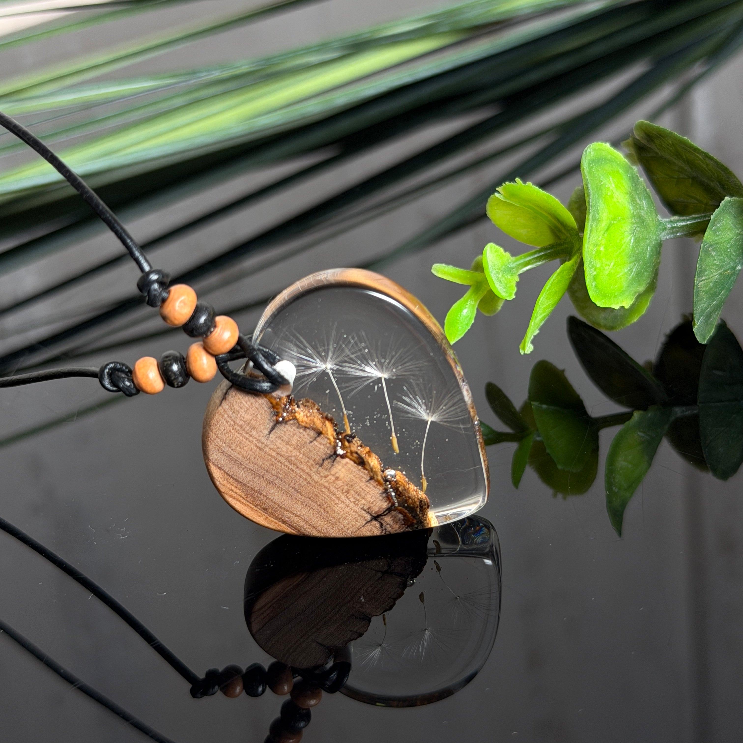 Handgefertigter Herzanhänger aus warm gemasertem Olivenholz und kristallklarem Epoxidharz mit schwebenden Pusteblumensamen. Schwarzes, verstellbares Lederband mit Schiebeknoten und naturfarbenen Holzperlen, Anhänger ca. 3,5 × 3,3 cm. Naturnaher Unikatschmuck von ZeitlosSchmuckDesign.