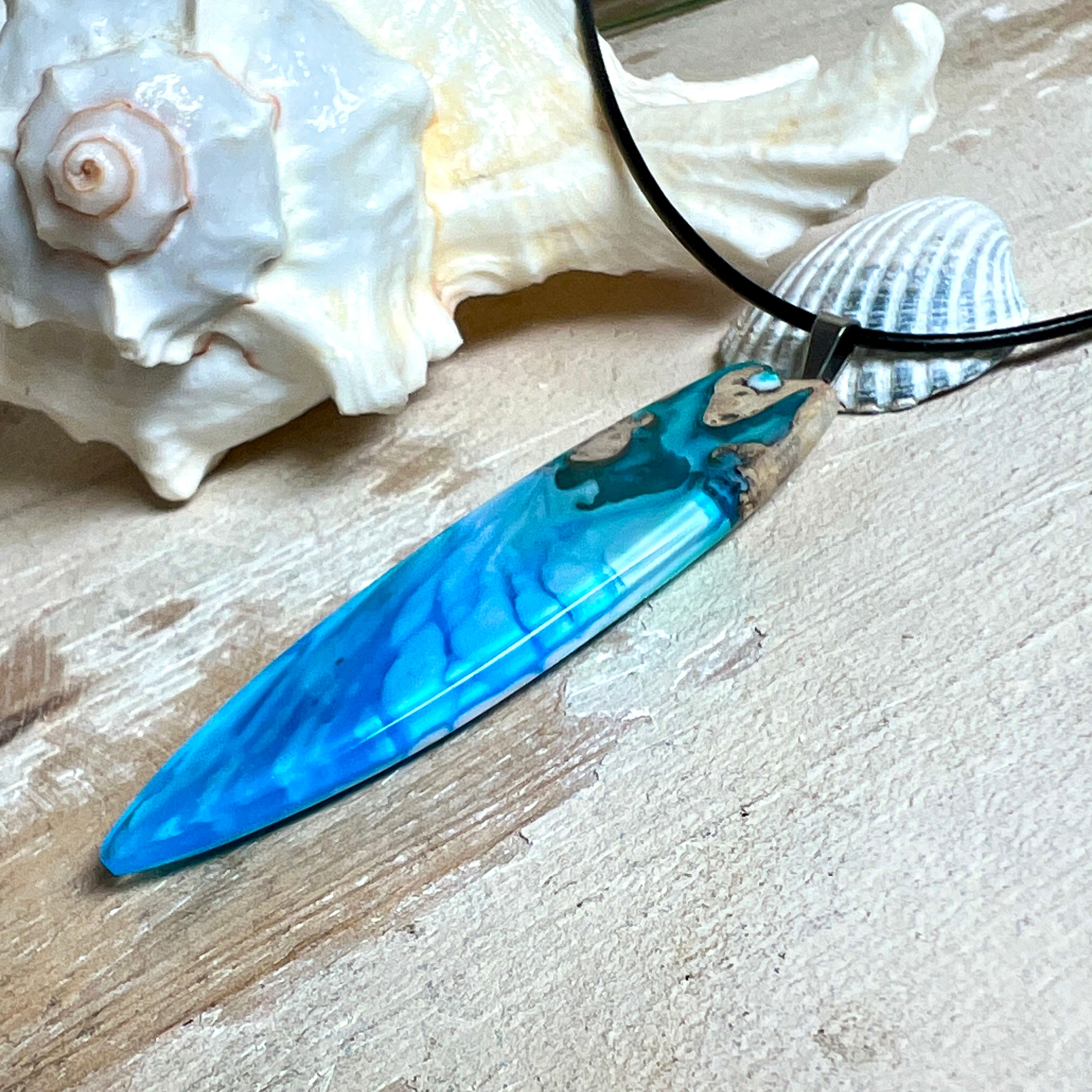 Surfbrett-Anhänger aus Maserholz und blau-weißem Epoxidharz, hochglanzpoliert, ca. 6,5 × 2 cm. An schwarzer Lederkette mit Edelstahl-Karabiner und 5-cm-Verlängerung; Edelstahl-Öse. Im Tageslicht lagunenblau, unter Schwarzlicht fluoreszierend – ideal als Party-/Festival-Schmuck. Handgefertigtes Unikat, Lieferung in edler Schmuckbox.