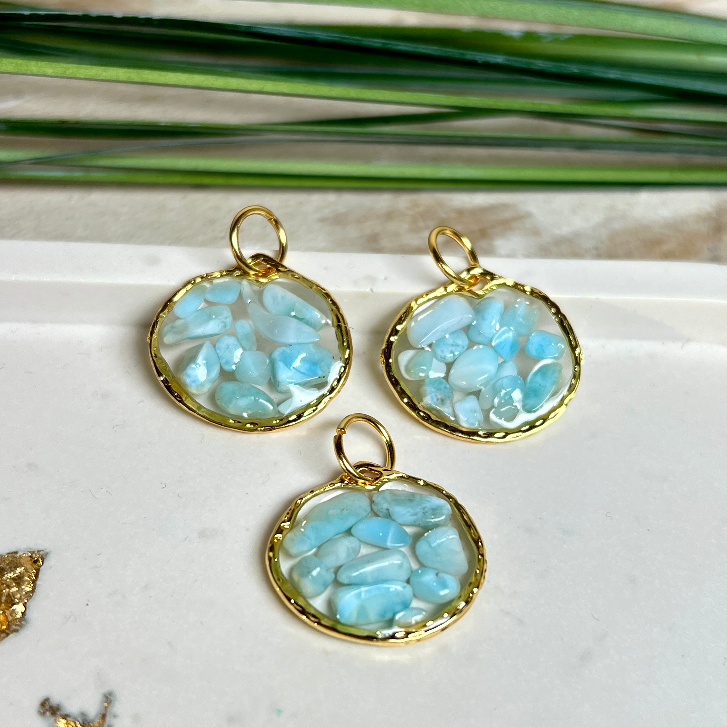 Runder Anhänger „Golden Atlantis Dream“ mit kleinen Larimar-Edelsteinchen in klarem Resin, gefasst in einer 24K vergoldeten Zinklegierung in Shiny Gold. Die Larimarsteine zeigen sanfte Blau- und Weißtöne wie karibische Wellen. Durchmesser ca. 22 mm, handgefertigtes Unikat, fotografiert mit Larimarsteinchen, Muscheln und einer 2-Euro-Münze zur Größenorientierung auf maritimem Untergrund.