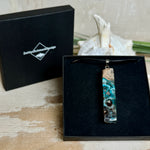 Rechteckiger Stabanhänger aus Maserholz und glasklarem Epoxidharz in tiefen Blau- und Türkistönen mit organischer Lagunen/Riff-Optik. Hochglanzpoliert, ca. 5,3 × 1,2 cm. Der Anhänger hängt an einer schwarzen Lederkette mit Edelstahl-Karabiner und 5-cm-Verlängerung (45 + 5 cm); die Anhängerschlaufe ist ebenfalls aus Edelstahl. Handgefertigtes Unikat, Lieferung in edler Schmuckbox – maritimer Look.