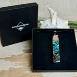 Rechteckiger Stabanhänger aus Maserholz und glasklarem Epoxidharz in tiefen Blau- und Türkistönen mit organischer Lagunen/Riff-Optik. Hochglanzpoliert, ca. 5,3 × 1,2 cm. Der Anhänger hängt an einer schwarzen Lederkette mit Edelstahl-Karabiner und 5-cm-Verlängerung (45 + 5 cm); die Anhängerschlaufe ist ebenfalls aus Edelstahl. Handgefertigtes Unikat, Lieferung in edler Schmuckbox – maritimer Look.