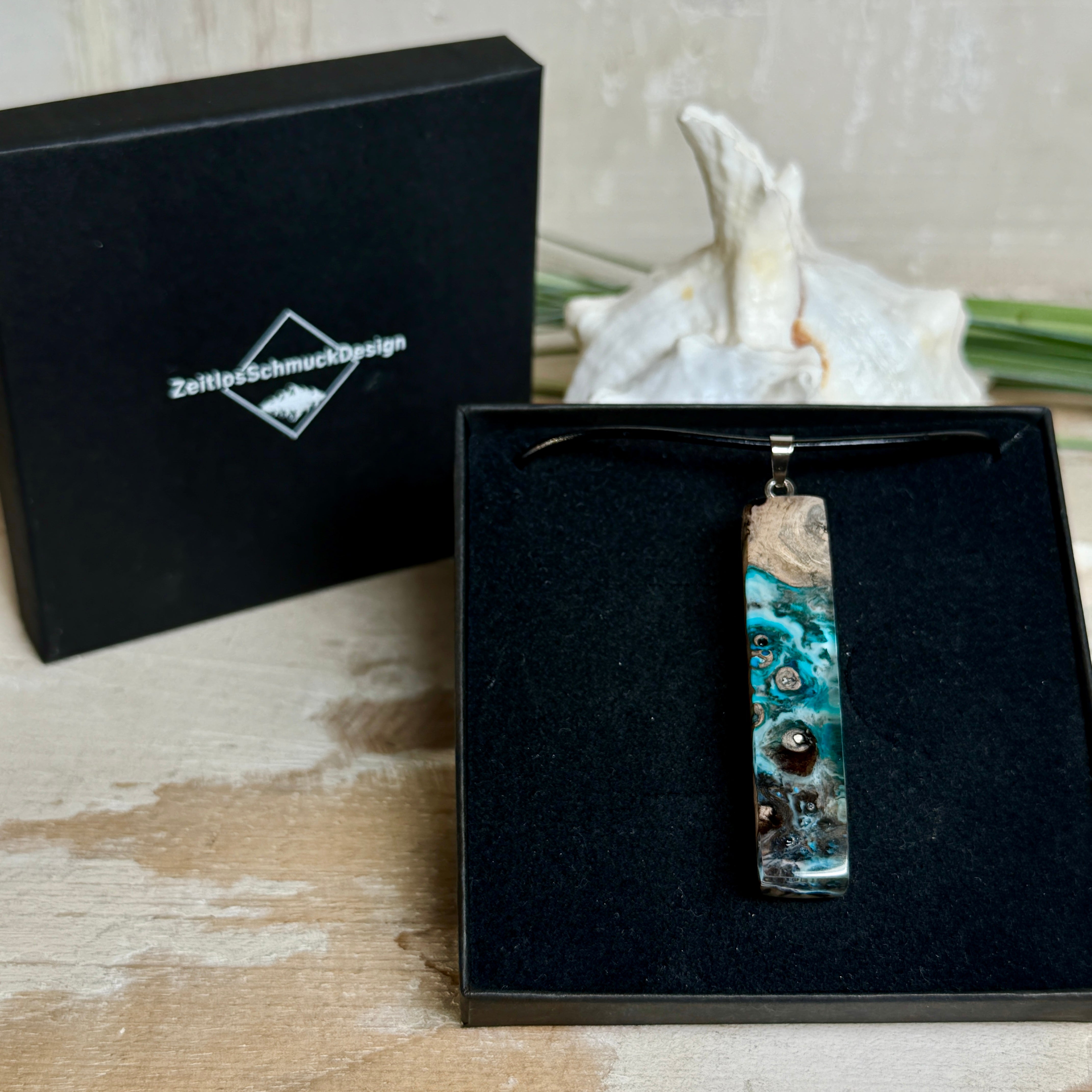 Rechteckiger Stabanhänger aus Maserholz und glasklarem Epoxidharz in tiefen Blau- und Türkistönen mit organischer Lagunen/Riff-Optik. Hochglanzpoliert, ca. 5,3 × 1,2 cm. Der Anhänger hängt an einer schwarzen Lederkette mit Edelstahl-Karabiner und 5-cm-Verlängerung (45 + 5 cm); die Anhängerschlaufe ist ebenfalls aus Edelstahl. Handgefertigtes Unikat, Lieferung in edler Schmuckbox – maritimer Look.