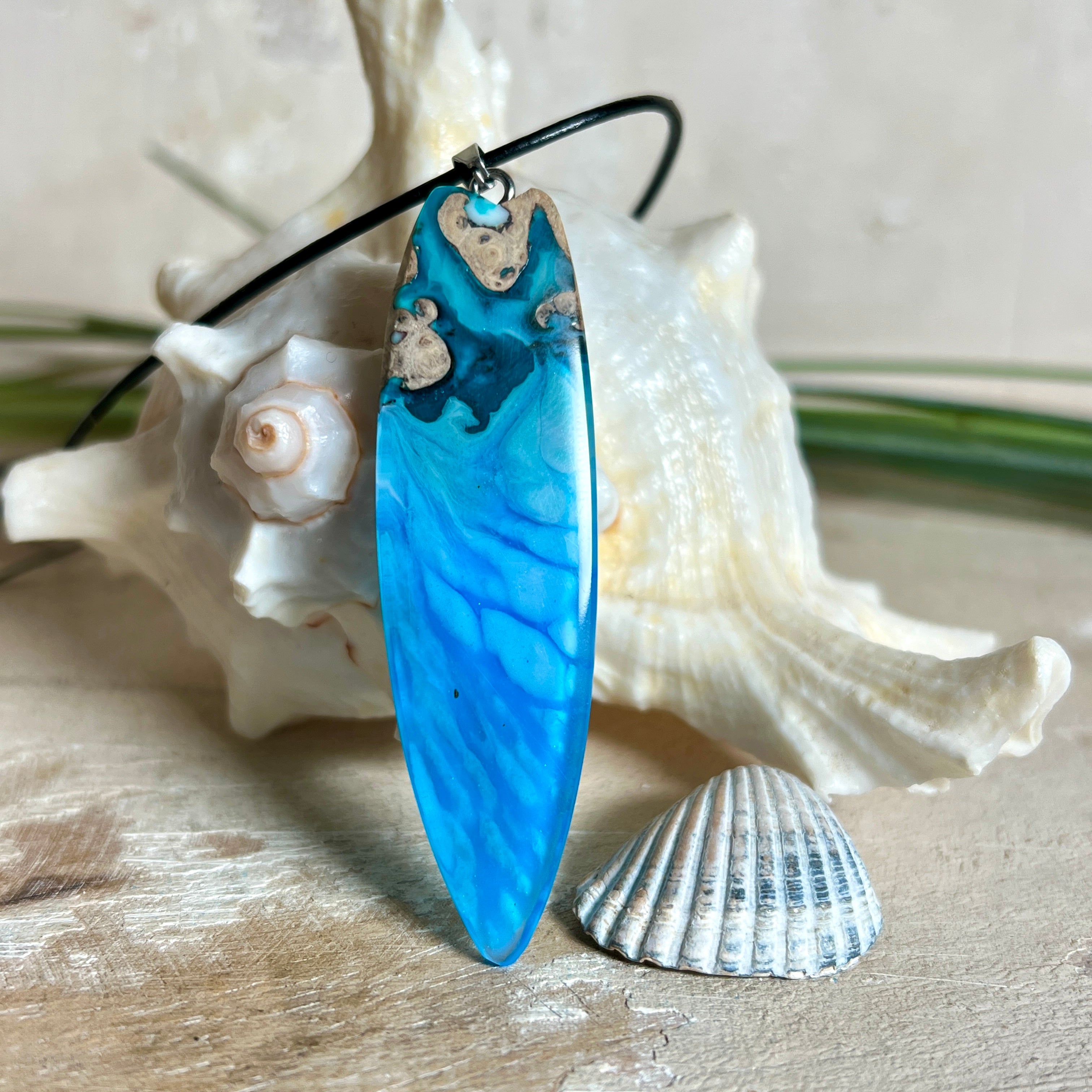 Surfbrett-Anhänger aus Maserholz und blau-weißem Epoxidharz, hochglanzpoliert, ca. 6,5 × 2 cm. An schwarzer Lederkette mit Edelstahl-Karabiner und 5-cm-Verlängerung; Edelstahl-Öse. Im Tageslicht lagunenblau, unter Schwarzlicht fluoreszierend – ideal als Party-/Festival-Schmuck. Handgefertigtes Unikat, Lieferung in edler Schmuckbox.