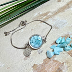 Verstellbares Edelstahlarmband mit Zirkoniasteinchen und Schiebeverschluss und rundem Anhänger (ca. 2,5 × 1,7 cm) mit echten Larimar-Edelstein-Splittern in Harz. Zusätzlich ein Charm „Blume des Lebens“ aus 925 Silber. Handgefertigtes Unikat in Türkis- und Meeresblautönen – Schmuckstück in Nahaufnahme bzw. als Tragebild am Handgelenk.