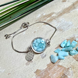 Verstellbares Edelstahlarmband mit Zirkoniasteinchen und Schiebeverschluss und rundem Anhänger (ca. 2,5 × 1,7 cm) mit echten Larimar-Edelstein-Splittern in Harz. Zusätzlich ein Charm „Blume des Lebens“ aus 925 Silber. Handgefertigtes Unikat in Türkis- und Meeresblautönen – Schmuckstück in Nahaufnahme bzw. als Tragebild am Handgelenk.