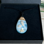 Handgefertigter Tropfenanhänger aus Maserholz und klarem Epoxidharz in Aquatönen. Im Harz sind echte Muscheln und ein kleiner Seestern eingebettet. Durch die besondere Mehrschicht-Gießtechnik wirkt es, als würden die Muscheln im Wasser schweben. Größe ca. 3,8 × 2 cm. Verstellbares Lederband (24–44 cm) mit Holzperlen. Maritimes Unikat.