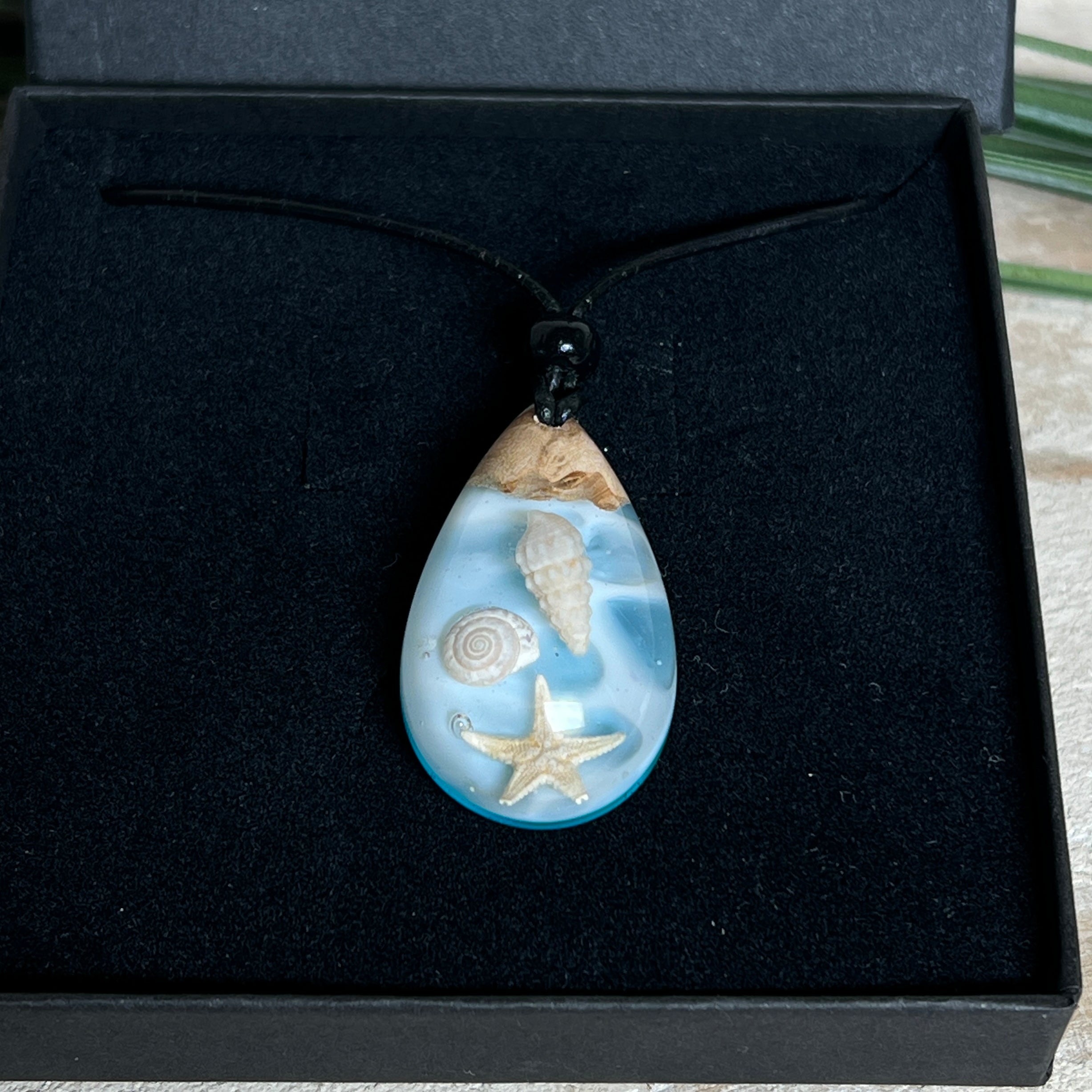 Handgefertigter Tropfenanhänger aus Maserholz und klarem Epoxidharz in Aquatönen. Im Harz sind echte Muscheln und ein kleiner Seestern eingebettet. Durch die besondere Mehrschicht-Gießtechnik wirkt es, als würden die Muscheln im Wasser schweben. Größe ca. 3,8 × 2 cm. Verstellbares Lederband (24–44 cm) mit Holzperlen. Maritimes Unikat.