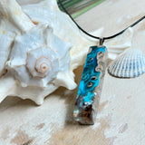 Rechteckiger Stabanhänger aus Maserholz und glasklarem Epoxidharz in tiefen Blau- und Türkistönen mit organischer Lagunen/Riff-Optik. Hochglanzpoliert, ca. 5,3 × 1,2 cm. Der Anhänger hängt an einer schwarzen Lederkette mit Edelstahl-Karabiner und 5-cm-Verlängerung (45 + 5 cm); die Anhängerschlaufe ist ebenfalls aus Edelstahl. Handgefertigtes Unikat, Lieferung in edler Schmuckbox – maritimer Look.