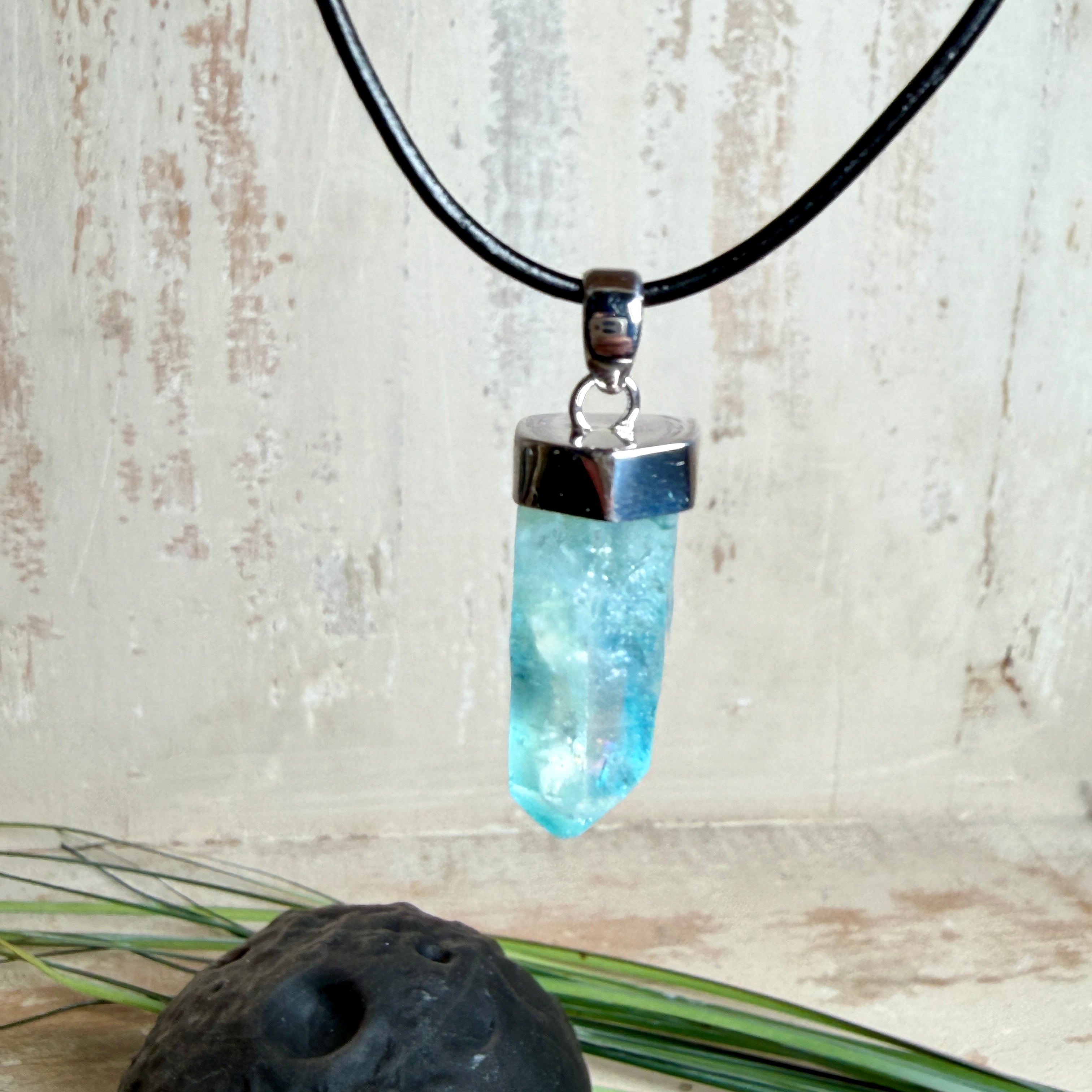 Nahaufnahme einer handgefertigten Halskette mit Aqua Aura Rohkristall: naturbelassener, sechskantiger Bergkristall mit goldbedampfter Aqua-Beschichtung in Türkisblau, gefasst in 925er Silber und getragen an einer schwarzen Lederkette mit Edelstahl-Karabiner und Verlängerungskettchen.