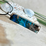 Rechteckiger Stabanhänger aus Maserholz und glasklarem Epoxidharz in tiefen Blau- und Türkistönen mit organischer Lagunen/Riff-Optik. Hochglanzpoliert, ca. 4,6 × 1,2 cm. Der Anhänger hängt an einer schwarzen Lederkette mit Edelstahl-Karabiner und 5-cm-Verlängerung (45 + 5 cm); die Anhängerschlaufe ist ebenfalls aus Edelstahl. Handgefertigtes Unikat, Lieferung in edler Schmuckbox – maritimer Look.