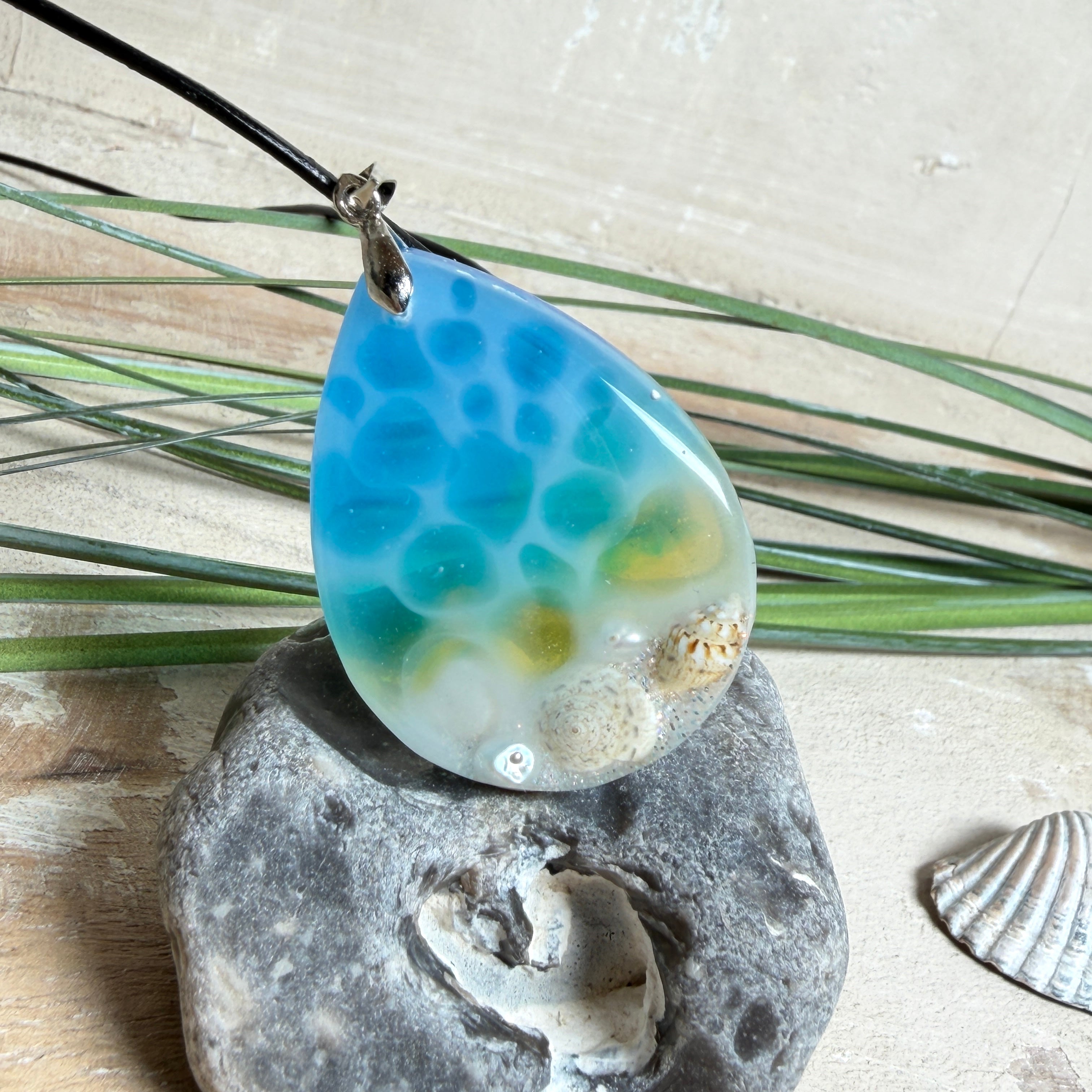 Tropfenförmiger Harzanhänger „Lagoon Drop“ in Blau-Grün-Weiß mit ca. 4 x 3 cm Größe. Im unteren Bereich sind kleine Muscheln, Sandstruktur und feine Süßwasserperlen eingegossen, sodass der Eindruck eines seichten Strandes im klaren Wasser entsteht. Der Anhänger hat eine Edelstahl-Schlaufe und hängt an einer schwarzen Lederkette mit Edelstahl-Karabiner und Verlängerung. Handgefertigter, maritime Unikat-Schmuck für Meeresliebhaberinnen.