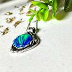 Handgemachter Anhänger mit rundem Cabochon aus Epoxidharz und schimmerndem synthetischem Opal. Die Farben changieren je nach Lichteinfall in Blau, Türkis und Grün. Der Opal sitzt in einer antik versilberten Fassung aus Messing (blei- & nickelfrei). Geliefert mit einer rhodinierten 925 Silberkette (45 cm). Jedes Stück ein Einzelstück – perfekt als stilvolles Geschenk oder persönlicher Glücksbringer.