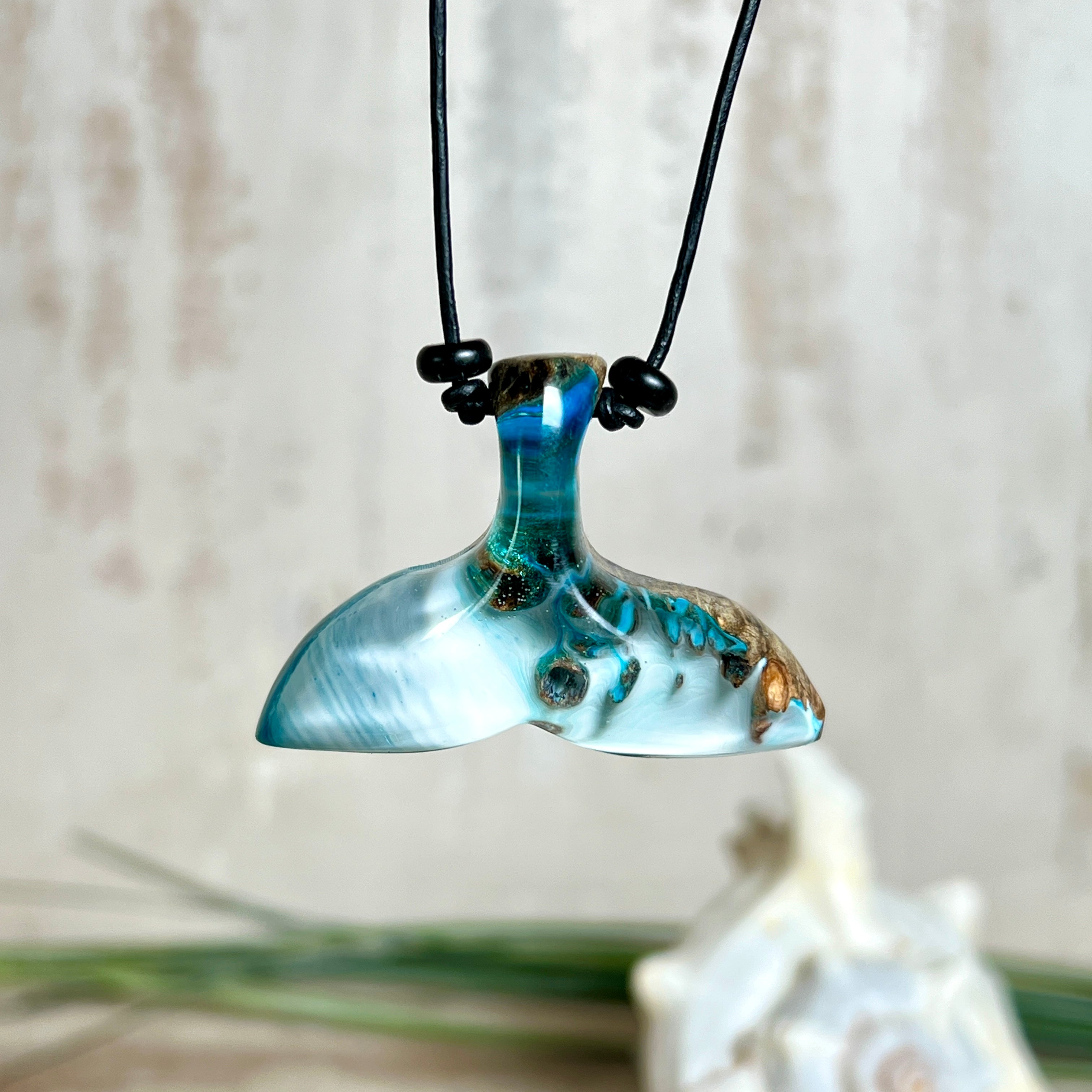 Handgefertigter Walflossen-Anhänger „Flossenklang“ aus Maserholz und türkis-blau-grau-weiß marmoriertem Epoxidharz an verstellbarem Lederband. Maritime Kette als Unikatschmuck für Meeresliebhaber und Wal-Fans, hochwertig poliert, präsentiert vor Muscheldeko und in Schmuckbox von ZeitlosSchmuckDesign.