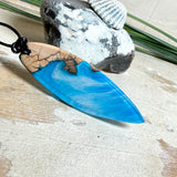 Handgefertigter Surfbrett-Anhänger aus warm gemasertem Olivenholz und Epoxidharz in Blau und Weiß, das an Meereswellen erinnert. Längliche, vorne polierte Form, ca. 6,2 × 2 cm, an verstellbarem schwarzem Lederband mit Knoten. Maritimer Unikatschmuck für Damen und Herren, dekorativ mit Stein und Muschel arrangiert.
