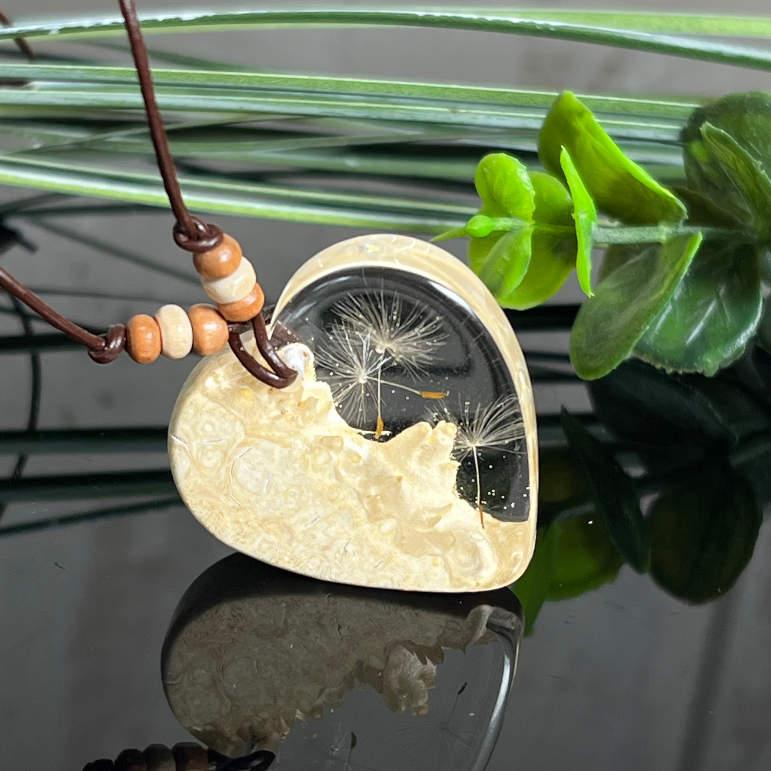 Herzanhänger aus Goldfield-Maserholz und transparentem Epoxidharz mit echter Pusteblume. Das handgefertigte Unikat kombiniert die natürliche Maserung des Holzes mit der zarten Leichtigkeit schwebender Pusteblumenschirmchen. Das braune Lederband mit Holzperlen ist verstellbar und verleiht dem Schmuckstück eine naturnahe, elegante Ausstrahlung.