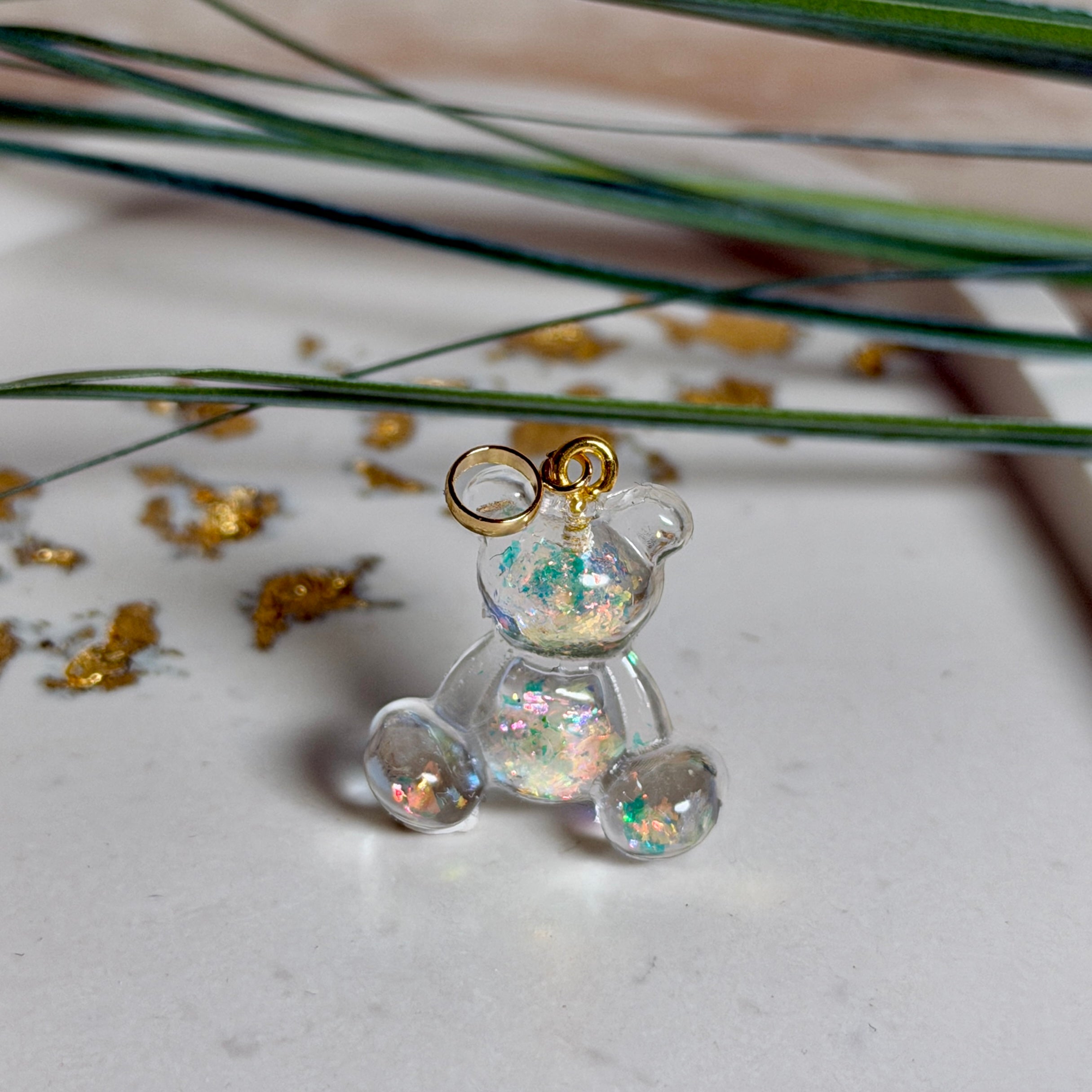 Kleiner Bärchen-Anhänger aus transparentem Harz mit irisierenden Flakes in Regenbogen-Schimmer, goldfarbene Anhängerschlaufe aus Edelstahl. Größe ca. 2,2 × 2,1 cm. Handgefertigtes Unikat; Flakes-Anordnung variiert, vereinzelt kleine Luftbläschen möglich.