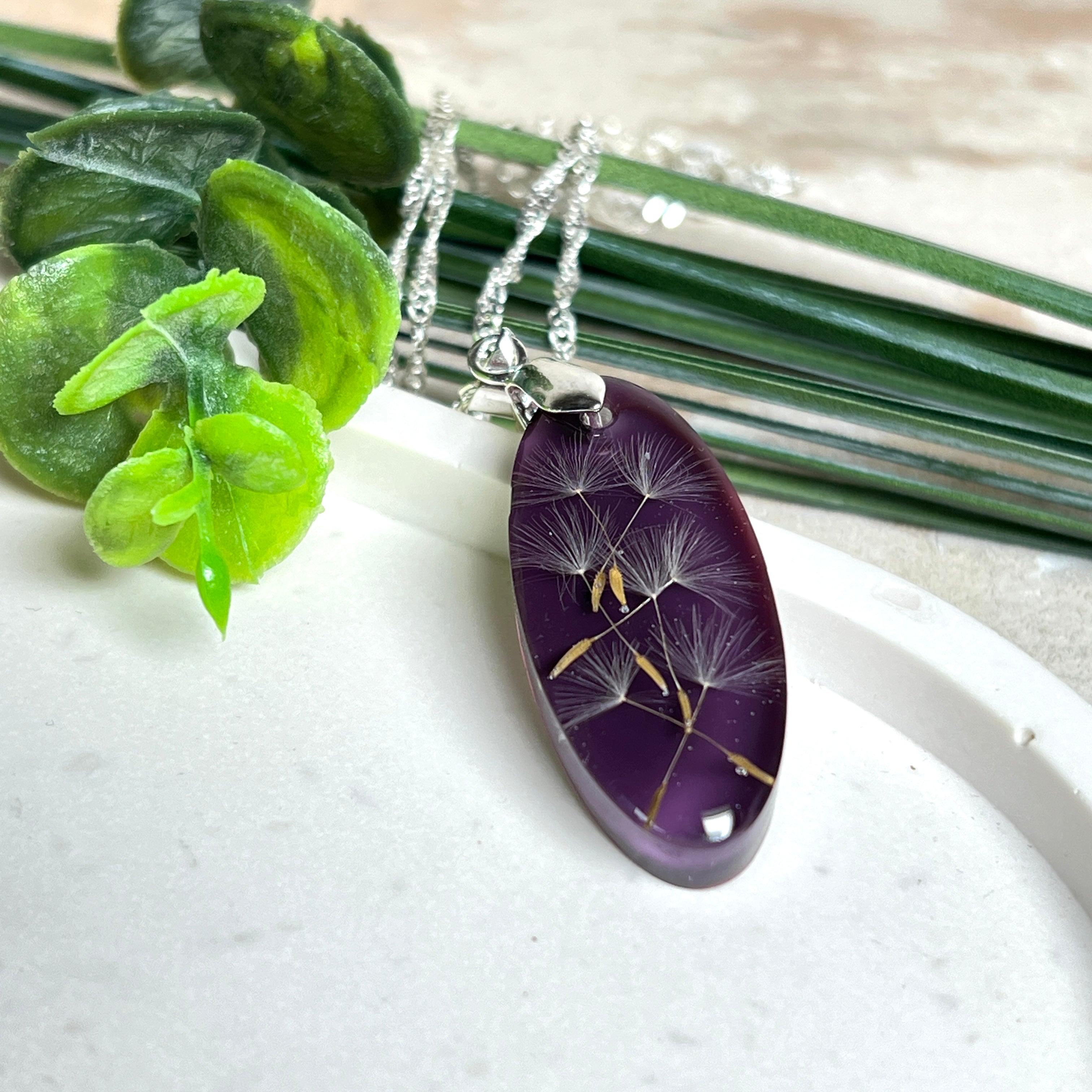 Ovaler Anhänger mit echter Pusteblume in Harz, Hintergrund Brombeerfarben. Größe 4 × 1,8 cm, versilberte Kette 50 cm mit Karabinerverschluss. Handgefertigtes Unikat.