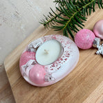 Rosa Teelichthalter aus klarem Epoxidharz, ca. 8 × 6,5 cm, Höhe ca. 4 cm. Im Harz sind kleine rosa Weihnachtskugeln, ein Stern, eine Schneeflocke und rosa Granulat eingegossen. Ovaler Halter für ein Teelicht, Unterseite mit vier runden Korkpads als Möbelschoner. Fotografiert auf Holz mit Tannenzweig und rosa Kugeln als weihnachtlicher Dekohintergrund.