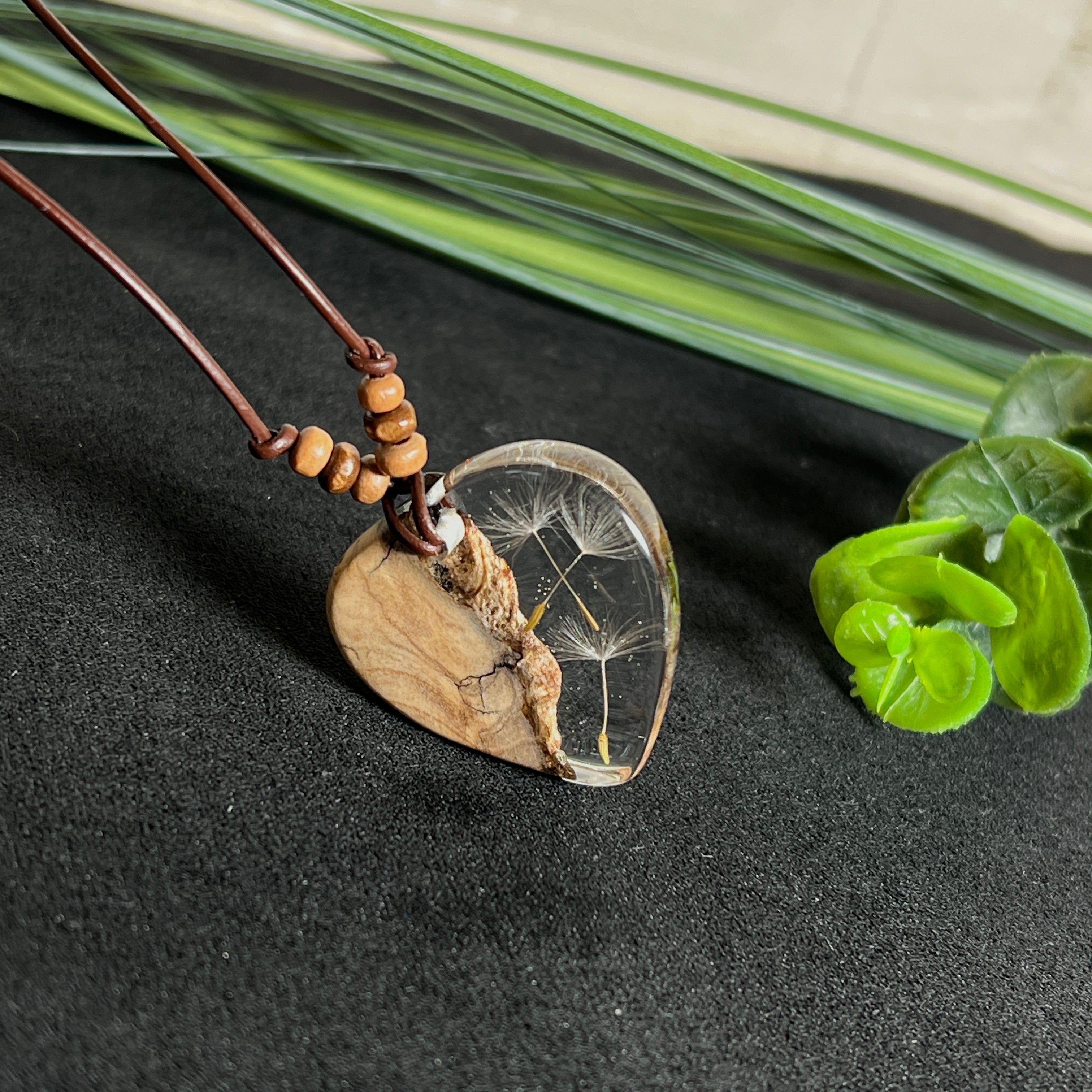 Handgefertigter Herzanhänger aus Olivenholz und klarem Harz mit echten Pusteblumen. Der Anhänger misst ca. 3,5 cm, das Lederband ist verstellbar von 23–43 cm und mit kleinen Holzperlen verziert. Ein natürliches Unikat, Symbol für Wünsche und Glück – sorgfältig gegossen, gesägt, geschliffen und poliert.