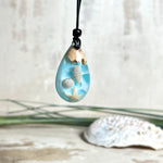 Handgefertigter Tropfenanhänger aus Maserholz und klarem Epoxidharz in Aquatönen. Im Harz sind echte Muscheln und ein kleiner Seestern eingebettet. Durch die besondere Mehrschicht-Gießtechnik wirkt es, als würden die Muscheln im Wasser schweben. Größe ca. 3,8 × 2 cm. Verstellbares Lederband (24–44 cm) mit Holzperlen. Maritimes Unikat.