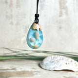 Handgefertigter Tropfenanhänger aus Maserholz und klarem Epoxidharz in Aquatönen. Im Harz sind echte Muscheln und ein kleiner Seestern eingebettet. Durch die besondere Mehrschicht-Gießtechnik wirkt es, als würden die Muscheln im Wasser schweben. Größe ca. 3,8 × 2 cm. Verstellbares Lederband (24–44 cm) mit Holzperlen. Maritimes Unikat.