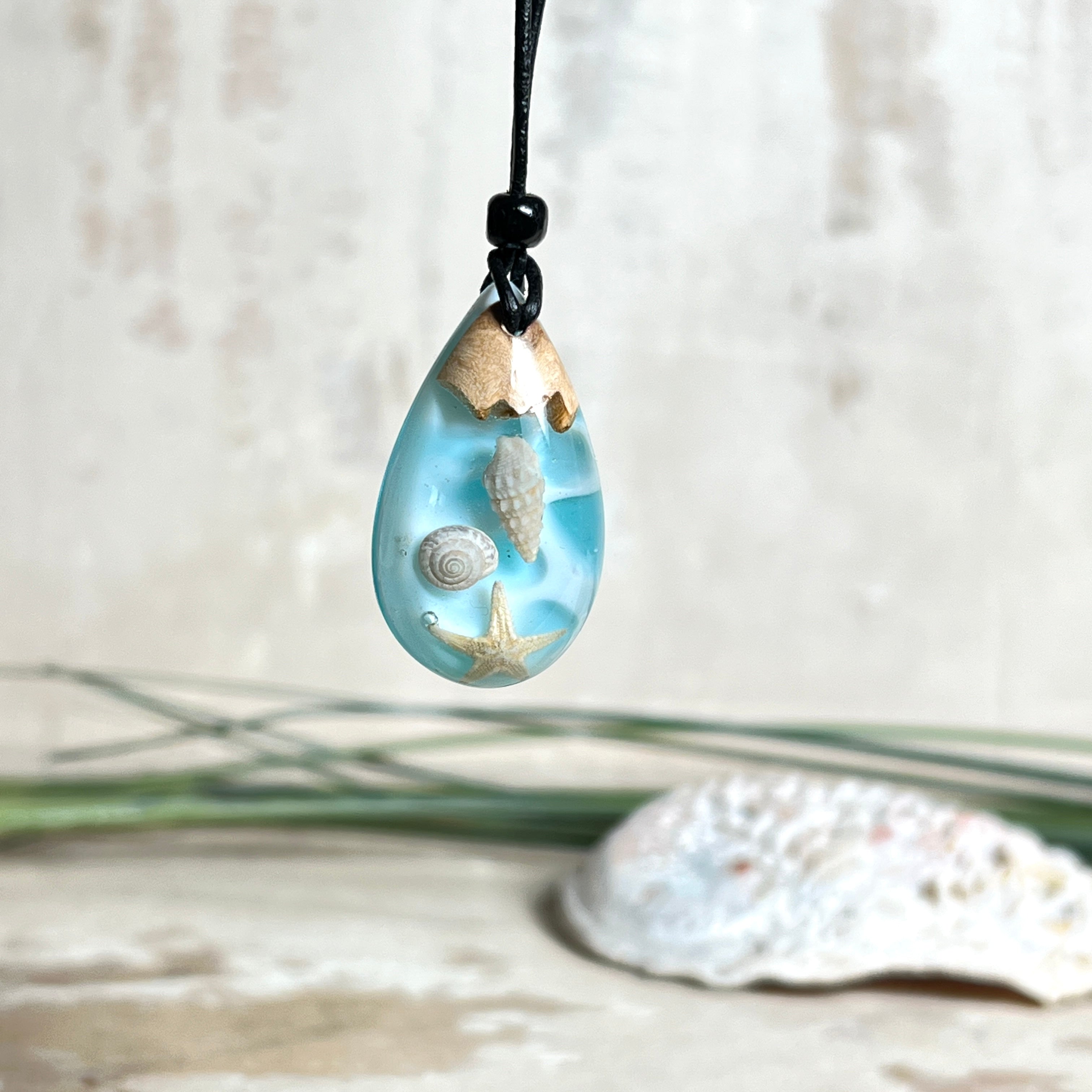 Handgefertigter Tropfenanhänger aus Maserholz und klarem Epoxidharz in Aquatönen. Im Harz sind echte Muscheln und ein kleiner Seestern eingebettet. Durch die besondere Mehrschicht-Gießtechnik wirkt es, als würden die Muscheln im Wasser schweben. Größe ca. 3,8 × 2 cm. Verstellbares Lederband (24–44 cm) mit Holzperlen. Maritimes Unikat.