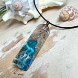 Rechteckiger Stabanhänger aus Maserholz und glasklarem Epoxidharz in tiefen Blau- und Türkistönen mit organischer Lagunen/Riff-Optik. Hochglanzpoliert, ca. 5,3 × 1,2 cm. Der Anhänger hängt an einer schwarzen Lederkette mit Edelstahl-Karabiner und 5-cm-Verlängerung (45 + 5 cm); die Anhängerschlaufe ist ebenfalls aus Edelstahl. Handgefertigtes Unikat, Lieferung in edler Schmuckbox – maritimer Look.