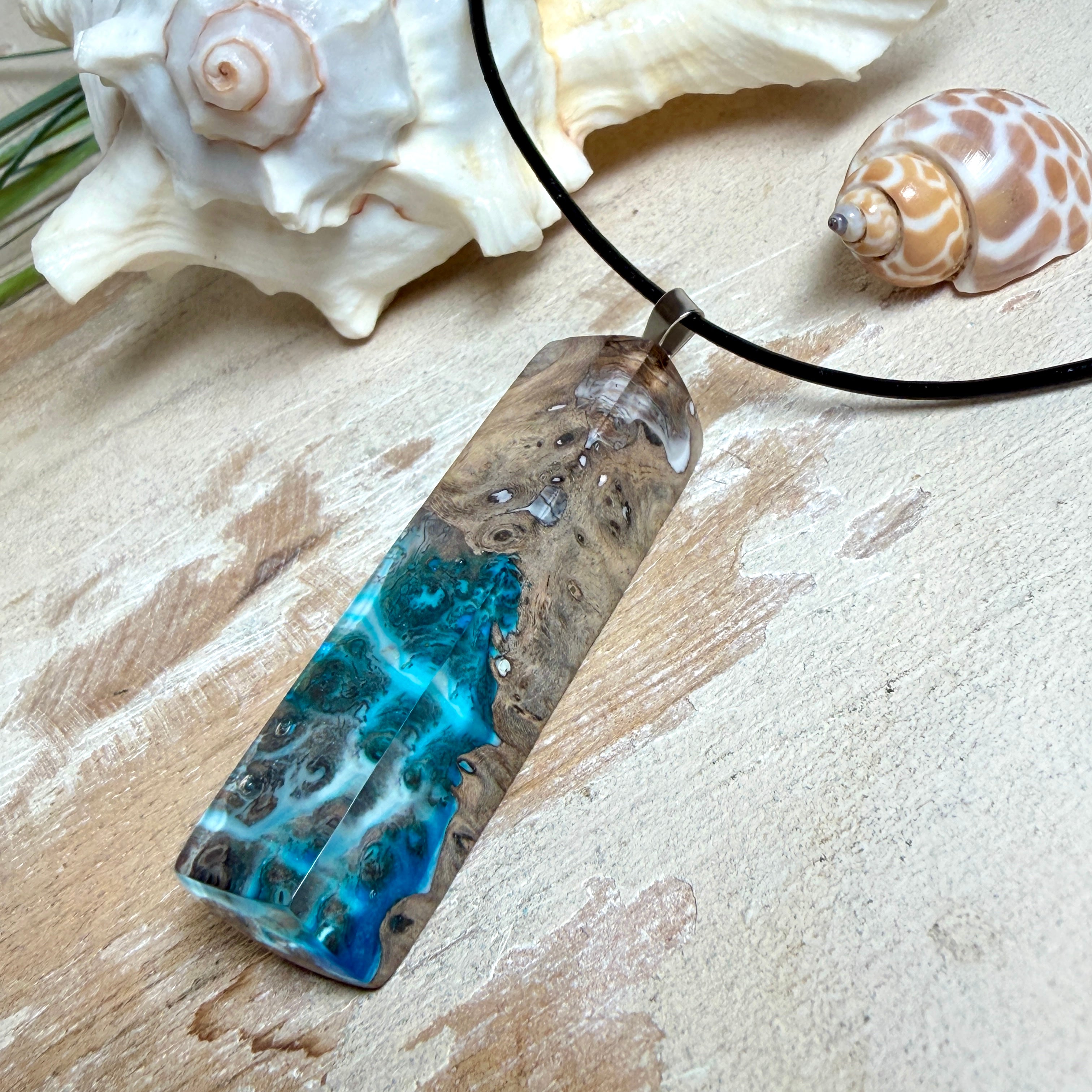 Rechteckiger Stabanhänger aus Maserholz und glasklarem Epoxidharz in tiefen Blau- und Türkistönen mit organischer Lagunen/Riff-Optik. Hochglanzpoliert, ca. 5,3 × 1,2 cm. Der Anhänger hängt an einer schwarzen Lederkette mit Edelstahl-Karabiner und 5-cm-Verlängerung (45 + 5 cm); die Anhängerschlaufe ist ebenfalls aus Edelstahl. Handgefertigtes Unikat, Lieferung in edler Schmuckbox – maritimer Look.