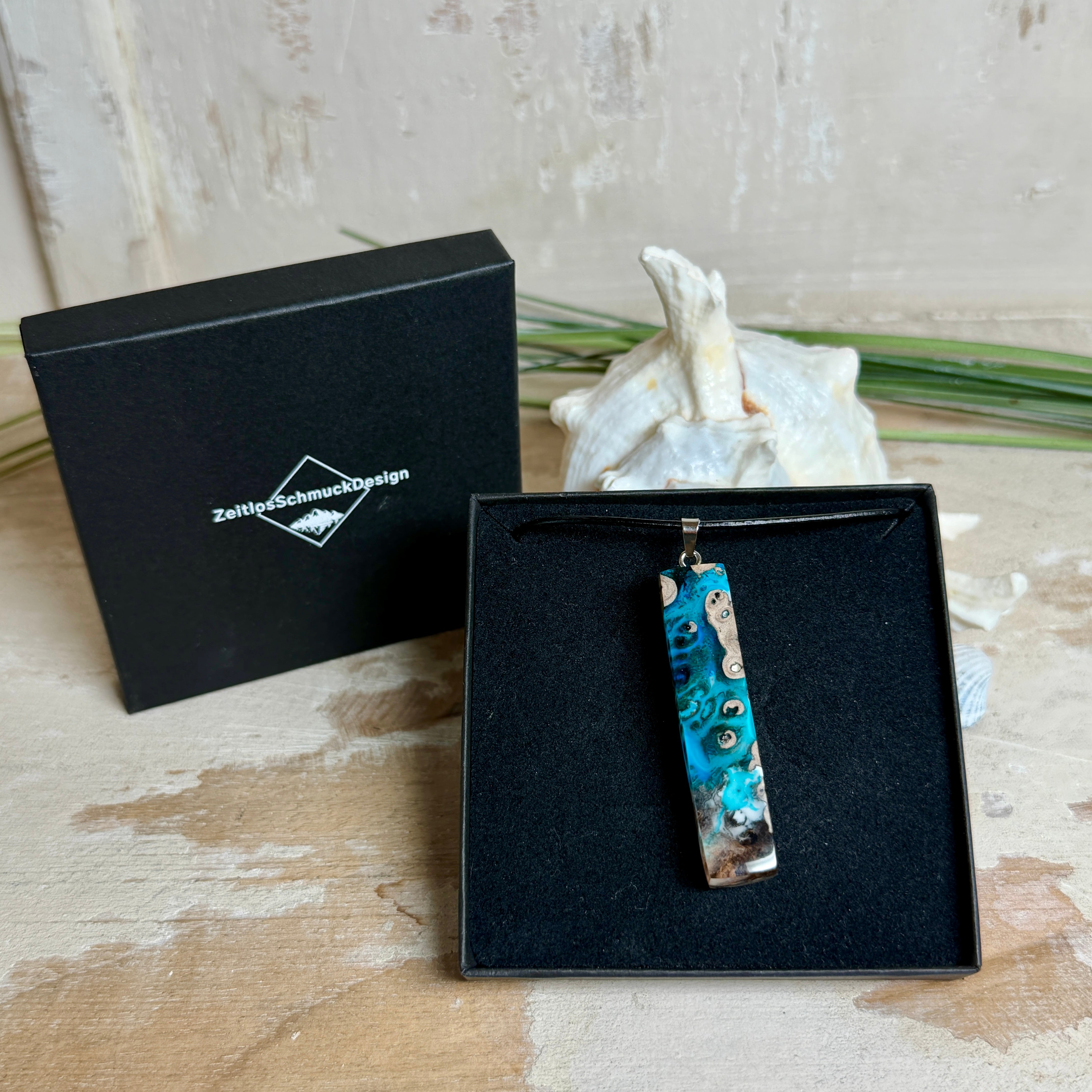 Rechteckiger Stabanhänger aus Maserholz und glasklarem Epoxidharz in tiefen Blau- und Türkistönen mit organischer Lagunen/Riff-Optik. Hochglanzpoliert, ca. 5,3 × 1,2 cm. Der Anhänger hängt an einer schwarzen Lederkette mit Edelstahl-Karabiner und 5-cm-Verlängerung (45 + 5 cm); die Anhängerschlaufe ist ebenfalls aus Edelstahl. Handgefertigtes Unikat, Lieferung in edler Schmuckbox – maritimer Look.