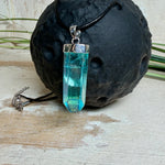 Nahaufnahme einer handgefertigten Halskette mit Aqua Aura Rohkristall: naturbelassener, sechskantiger Bergkristall mit goldbedampfter Aqua-Beschichtung in Türkisblau, gefasst in 925er Silber und getragen an einer schwarzen Lederkette mit Edelstahl-Karabiner und Verlängerungskettchen.