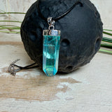 Nahaufnahme einer handgefertigten Halskette mit Aqua Aura Rohkristall: naturbelassener, sechskantiger Bergkristall mit goldbedampfter Aqua-Beschichtung in Türkisblau, gefasst in 925er Silber und getragen an einer schwarzen Lederkette mit Edelstahl-Karabiner und Verlängerungskettchen.