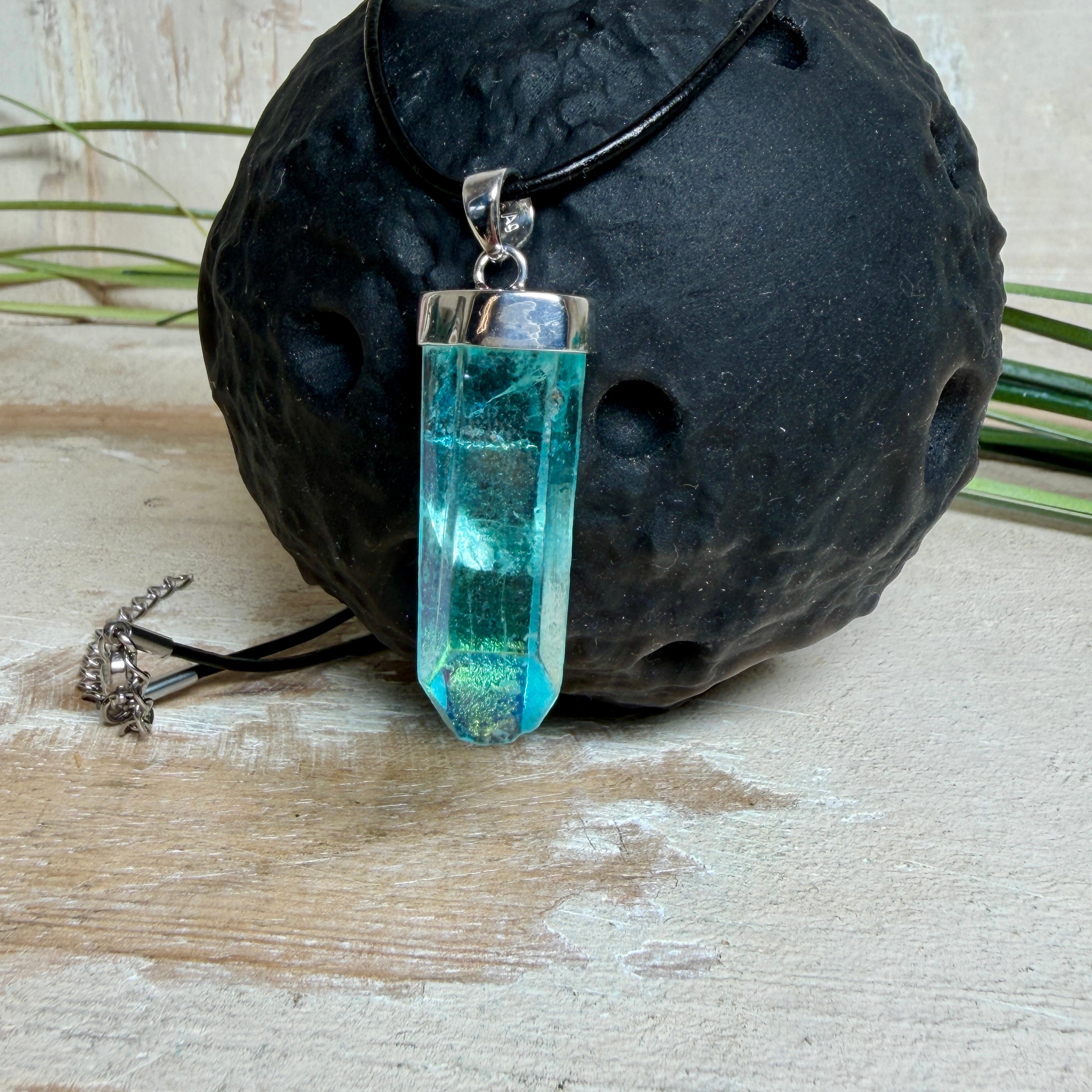 Nahaufnahme einer handgefertigten Halskette mit Aqua Aura Rohkristall: naturbelassener, sechskantiger Bergkristall mit goldbedampfter Aqua-Beschichtung in Türkisblau, gefasst in 925er Silber und getragen an einer schwarzen Lederkette mit Edelstahl-Karabiner und Verlängerungskettchen.