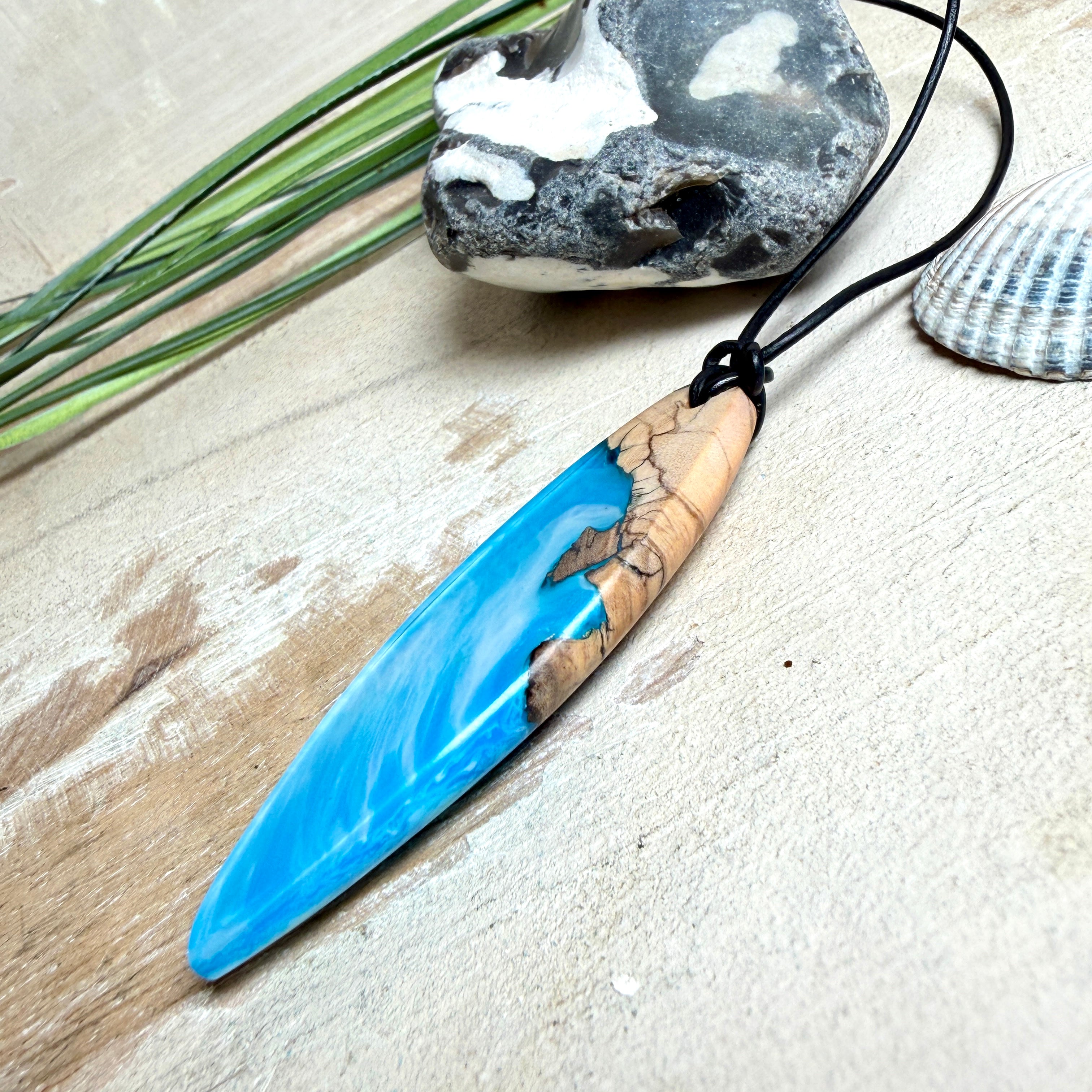 Handgefertigter Surfbrett-Anhänger aus warm gemasertem Olivenholz und Epoxidharz in Blau und Weiß, das an Meereswellen erinnert. Längliche, vorne polierte Form, ca. 6,2 × 2 cm, an verstellbarem schwarzem Lederband mit Knoten. Maritimer Unikatschmuck für Damen und Herren, dekorativ mit Stein und Muschel arrangiert.