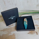 Unikat Anhänger aus stabilisiertem Holz mit türkisblauem Harzverlauf, schlanke Form, poliert. Verstellbares braunes Lederband 22–46 cm mit Schiebeknoten. Anhänger ca. 6,6 × 2 cm. Tragefoto am Hals und Produktfotos mit maritimer Deko