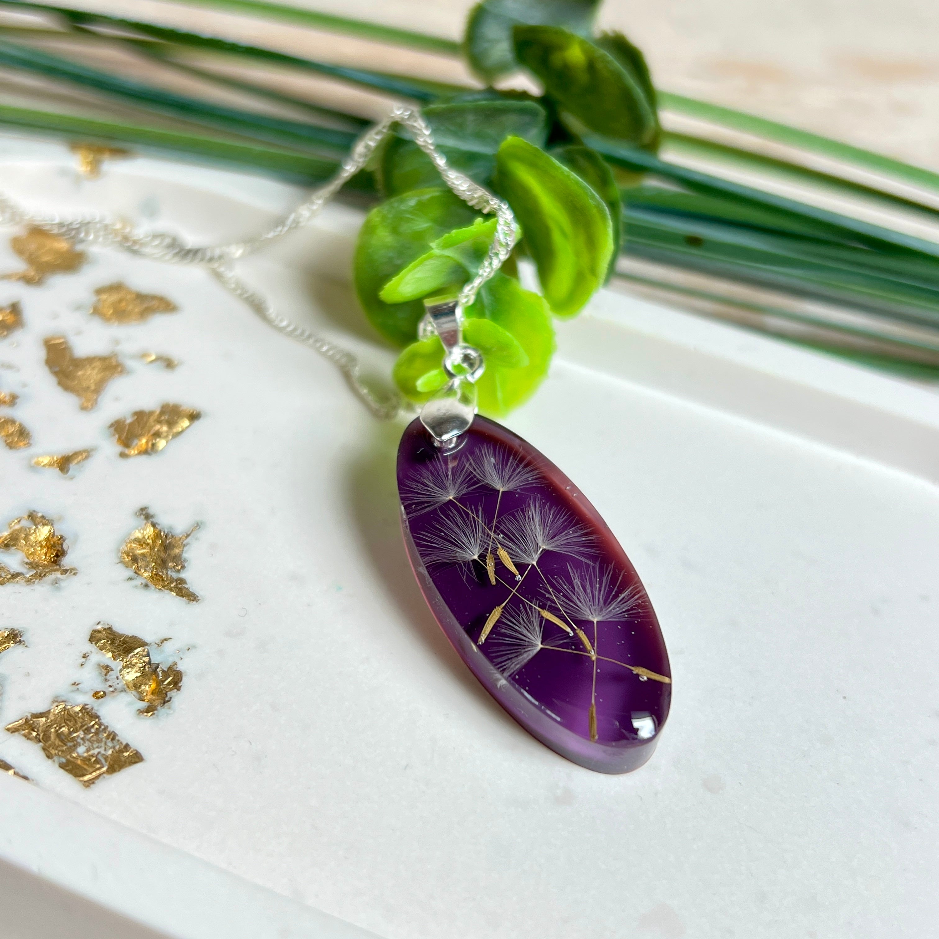 Ovaler Anhänger mit echter Pusteblume in Harz, Hintergrund Brombeerfarben. Größe 4 × 1,8 cm, versilberte Kette 50 cm mit Karabinerverschluss. Handgefertigtes Unikat.