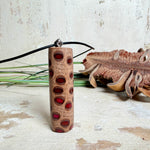 Handgefertigter Halsanhänger aus echtem Banksiazapfen, veredelt mit rotem Harz in den natürlichen Kammern; ca. 5,5 × 1,6 cm. Mit Anhängerschlaufe aus Edelstahl an schwarzer Lederkette (45 cm + 5 cm Verlängerung) mit Edelstahl-Karabiner. Detailaufnahme bzw. Tragebild vor neutralem, natürlichem Hintergrund.