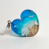 Ein herzförmiger Anhänger mit einer ganzen Ozeanwelt darin: Welle, Sand und Seestern in leuchtendem Blau und Türkis – handgegossen, einzigartig, unvergesslich. Mit versilberter Kette (46 cm) und Edelstahl-Karabiner.