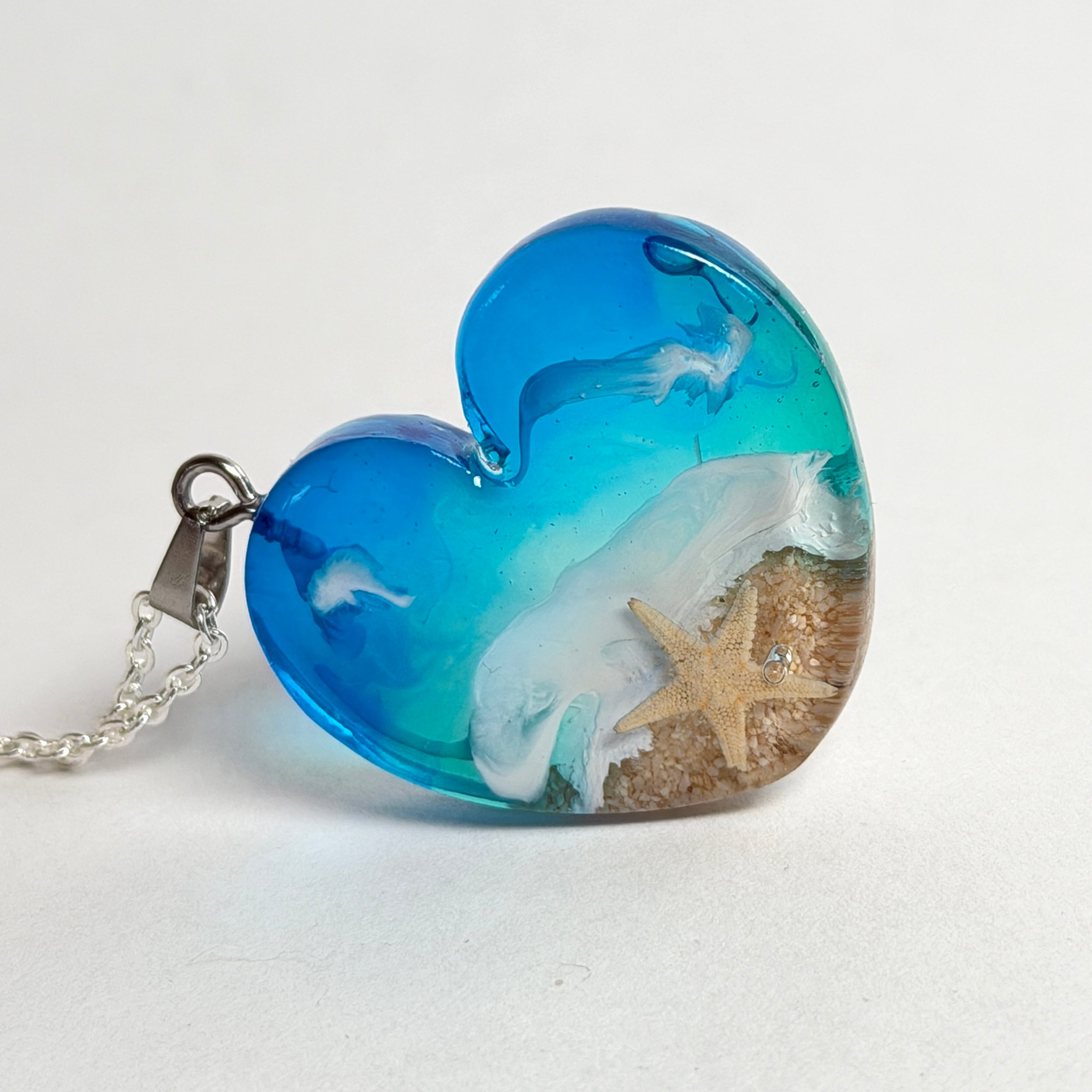Ein herzförmiger Anhänger mit einer ganzen Ozeanwelt darin: Welle, Sand und Seestern in leuchtendem Blau und Türkis – handgegossen, einzigartig, unvergesslich. Mit versilberter Kette (46 cm) und Edelstahl-Karabiner.