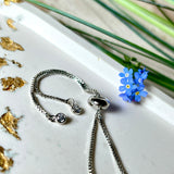 „Detailaufnahme eines handgefertigten Armbands mit echter blauer Vergissmeinnicht und feinen gelben Blüten in Harz, eingefasst in sechseckiger versilberter Lünette. Verstellbares Edelstahlarmband mit funkelnden Zirkonia-Endperlen – liebevolles Natur-Schmuckstück, ideal als Geschenk für Frauen.“