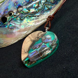 Herzförmiger Anhänger aus Olivenholz mit aquamarinfarbenem Harz und eingelegten Paua-Muschelstücken (Abalone), irisierender Blau-Grün-Schimmer. Der Anhänger hängt an einem braunen, verstellbaren Lederband mit Schiebeknoten; Aufhängung aus Edelstahl. Produktfoto vor maritimer Deko/Abalone-Schale.