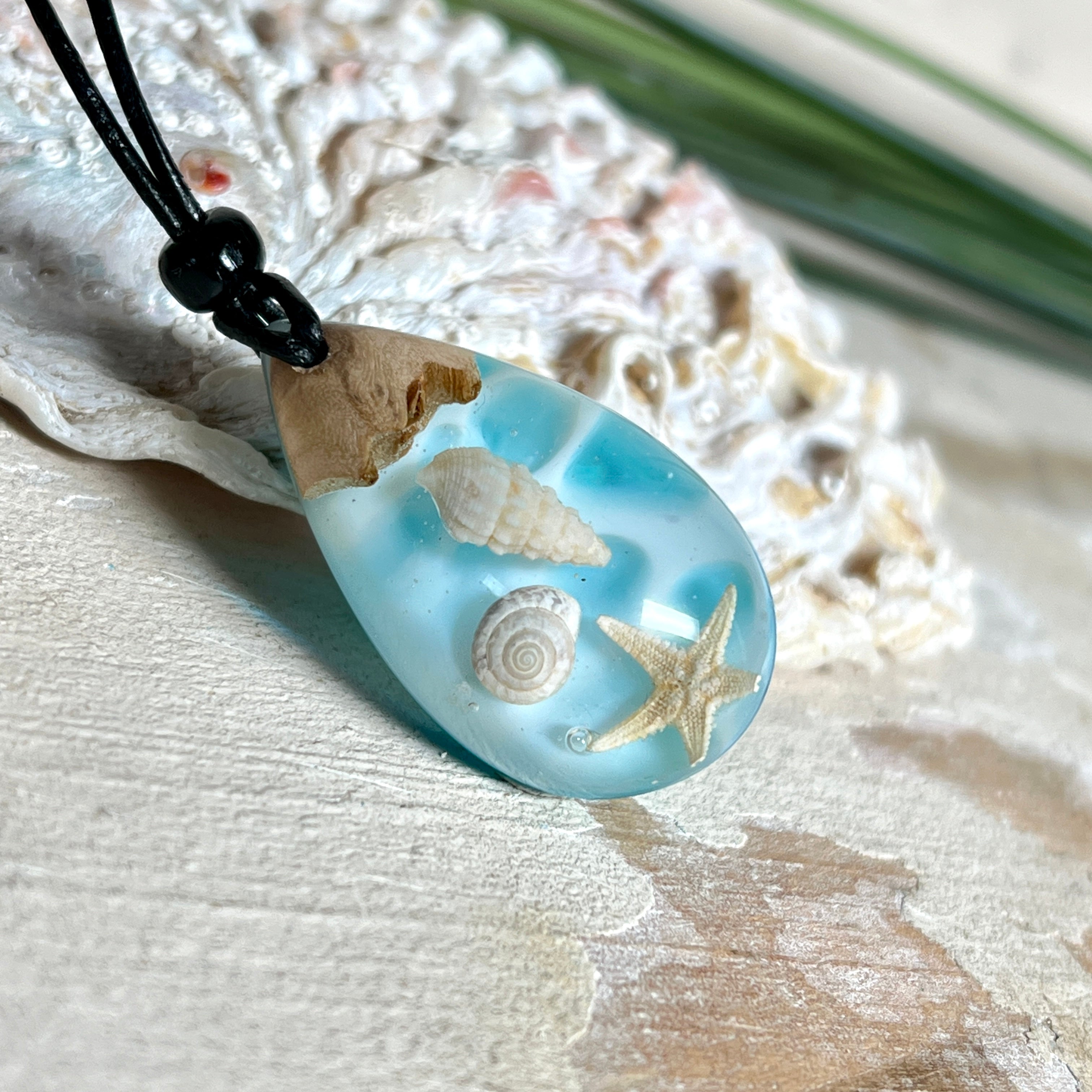 Handgefertigter Tropfenanhänger aus Maserholz und klarem Epoxidharz in Aquatönen. Im Harz sind echte Muscheln und ein kleiner Seestern eingebettet. Durch die besondere Mehrschicht-Gießtechnik wirkt es, als würden die Muscheln im Wasser schweben. Größe ca. 3,8 × 2 cm. Verstellbares Lederband (24–44 cm) mit Holzperlen. Maritimes Unikat.